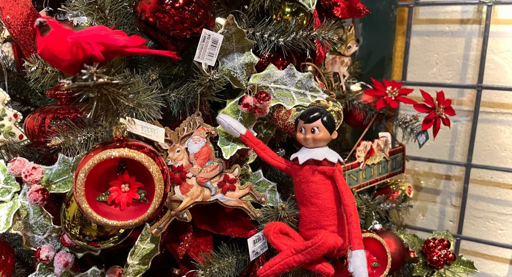 An elf on the shelf sits in the tree helping to decorate with a Santa ornament.